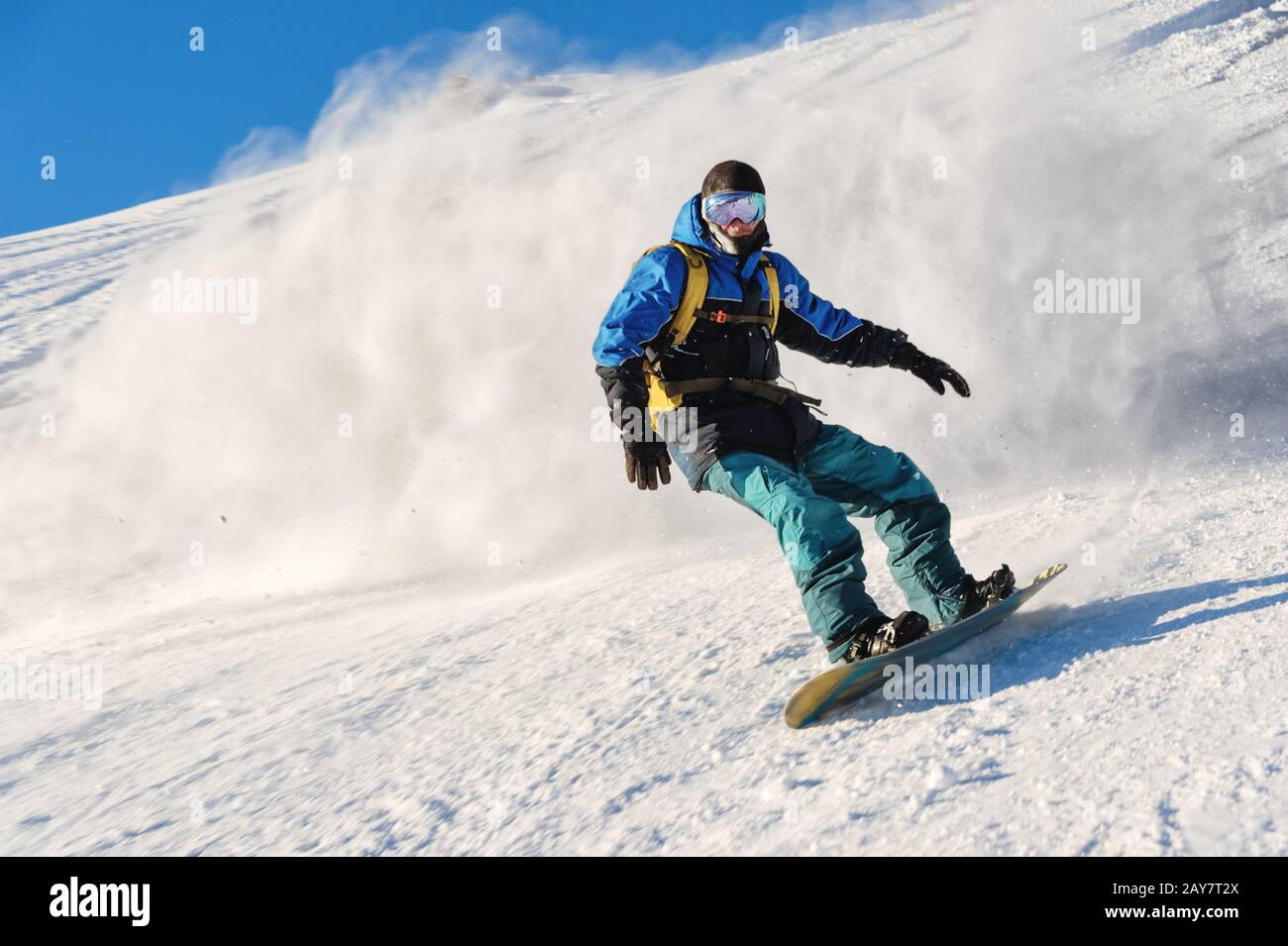 Freeride Snowboarder rollt auf einer schneebedeckten Piste, die hinter einem Schneepulver gegen den blauen Himmel zurückbleibt Stockfoto