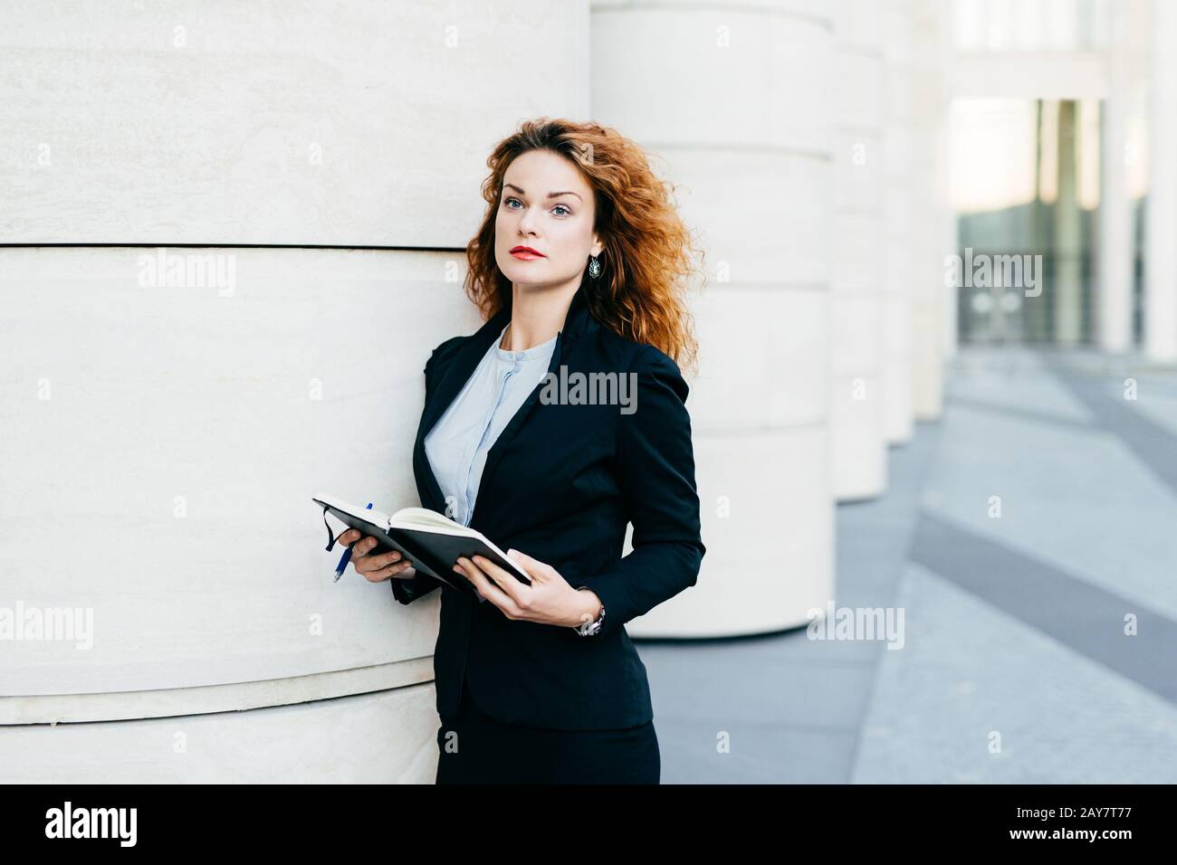Hübsche junge Geschäftsfrau, die schwarze Jacke, Rock und weiße Bluse trägt, ihr Taschenbuch mit Stift hält, Notizen schreibt oder nach Ersatztagen sucht Stockfoto