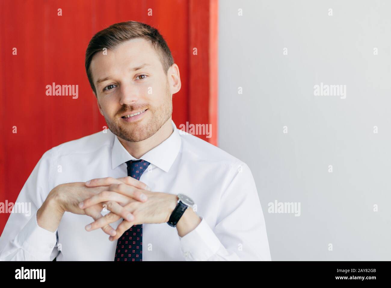 Foto eines gutaussehenden intelligenten Mannes mit sanftem Lächeln, hält die Hände zusammen, trägt formales weißes Hemd, posiert vor rot-weißem Hintergrund. Männliche Busi Stockfoto