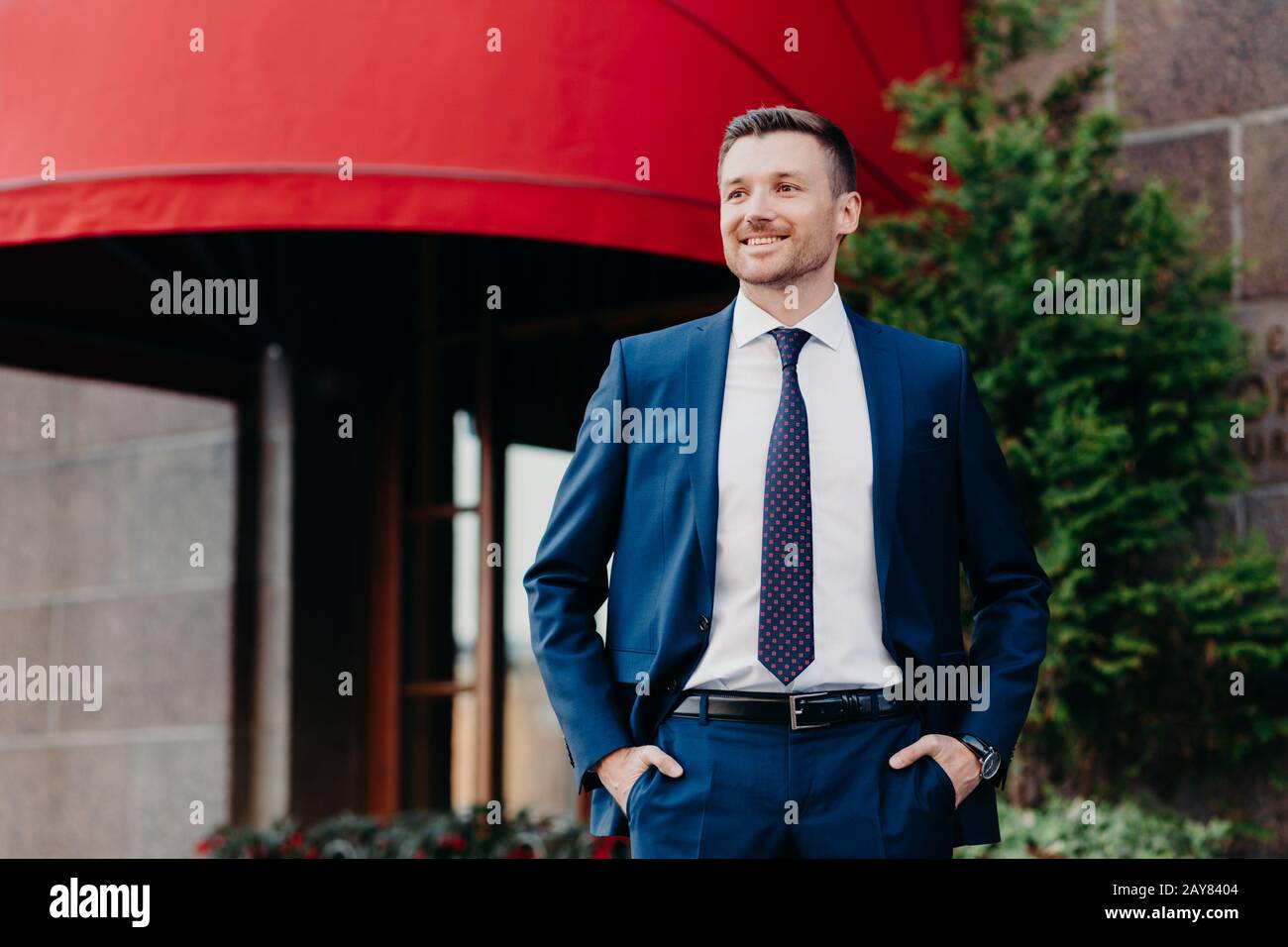 Der attraktive junge Unternehmer sieht selbstbewusst und glücklich vor der Kamera aus, hält die Hände in den Taschen, trägt teuren Anzug, hat ein positives Lächeln, ist zufrieden Stockfoto