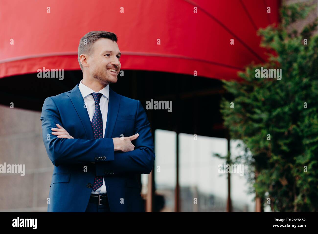 Froh wohlhabender Geschäftsmann hält Arm gefaltet, trägt formellen Anzug, wirkt mit positivem Ausdruck beiseite, freut sich über Erfolg und hohen Umsatz, posiert draußen Stockfoto