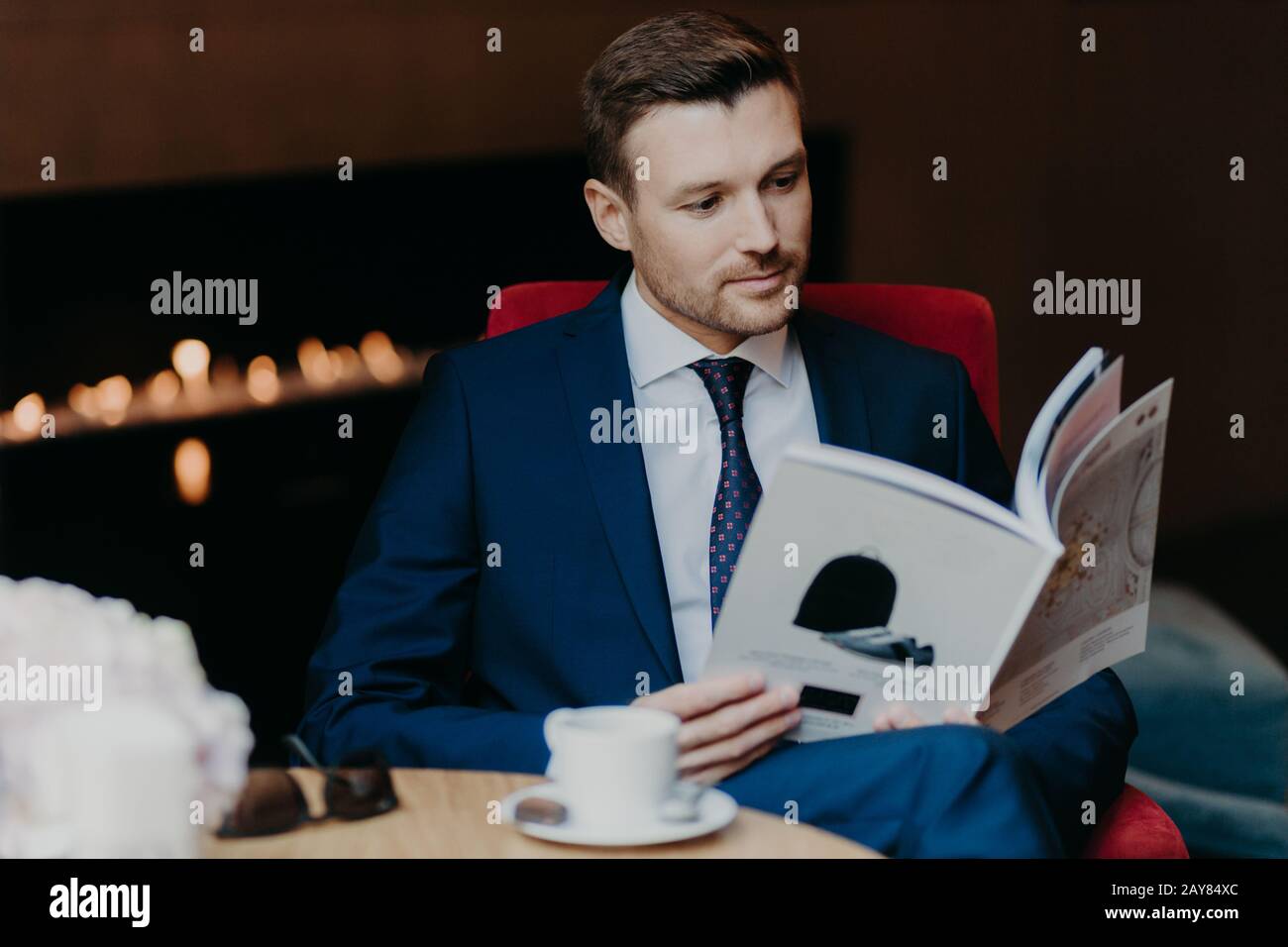 Der seriöse männliche Unternehmer liest das Wirtschaftsmagazin, sitzt im Café, trinkt Kaffee oder Tee, ist in einem formellen Anzug gekleidet, hat eine Pause zum Abendessen und ruht danach Stockfoto
