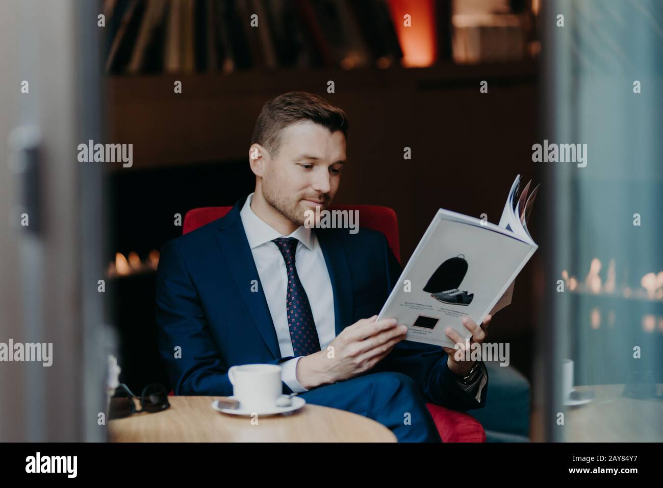 Gutaussehender junger Geschäftsmann schaut aufmerksam in die Zeitschrift, gekleidet in formellen Anzug, trinkt aromatischen Kaffee oder Cappuccino, genießt Dinnerpause, ruhiger Armo Stockfoto