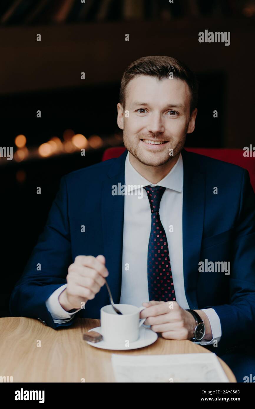 Zufriedener männlicher Mitarbeiter im Luxusanzug, Kaffee trinken, sitzt am Holztisch in der Cafeteria, hat ein angenehmes Lächeln und einen attraktiven Look, wartet auf Business p Stockfoto