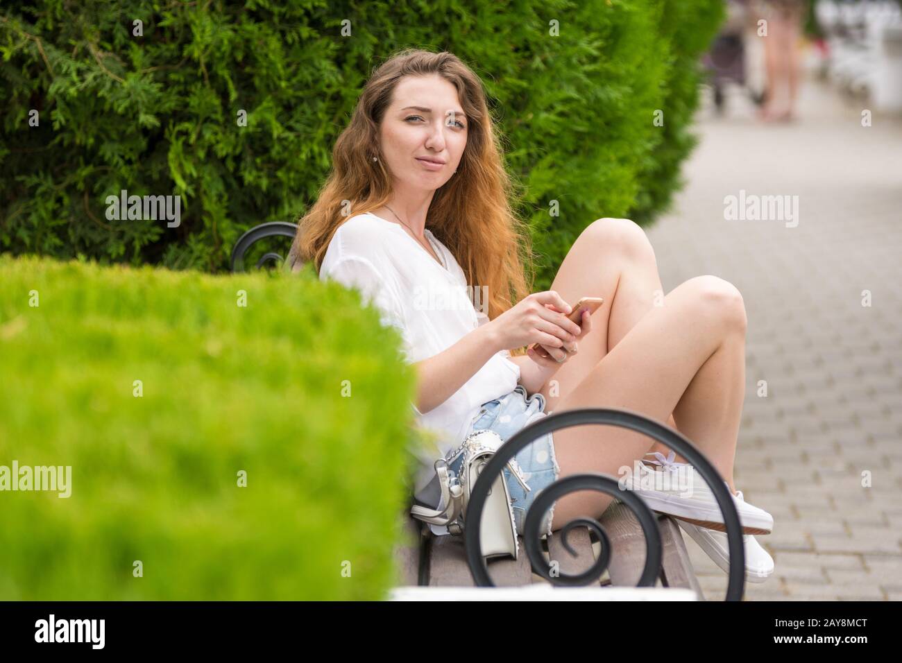 Ein junges Mädchen mit einem Telefon in der Hand, sitzt auf einer Bank im Park und blickte auf den Rahmen Stockfoto