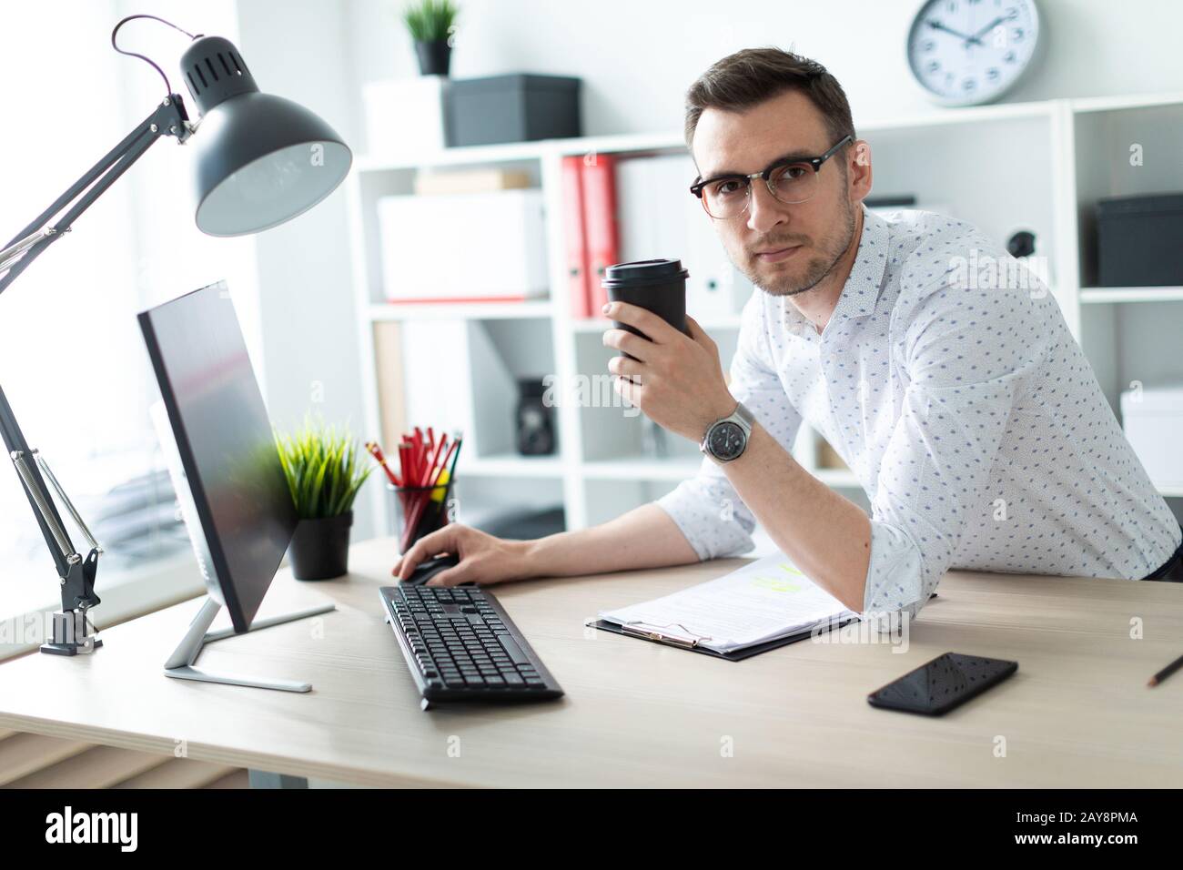 Ein junger Mann in Gläsern steht in der Nähe eine Tabelle im Büro, hält ein Glas Kaffee in der Hand und mit einem Computer funktioniert. Stockfoto
