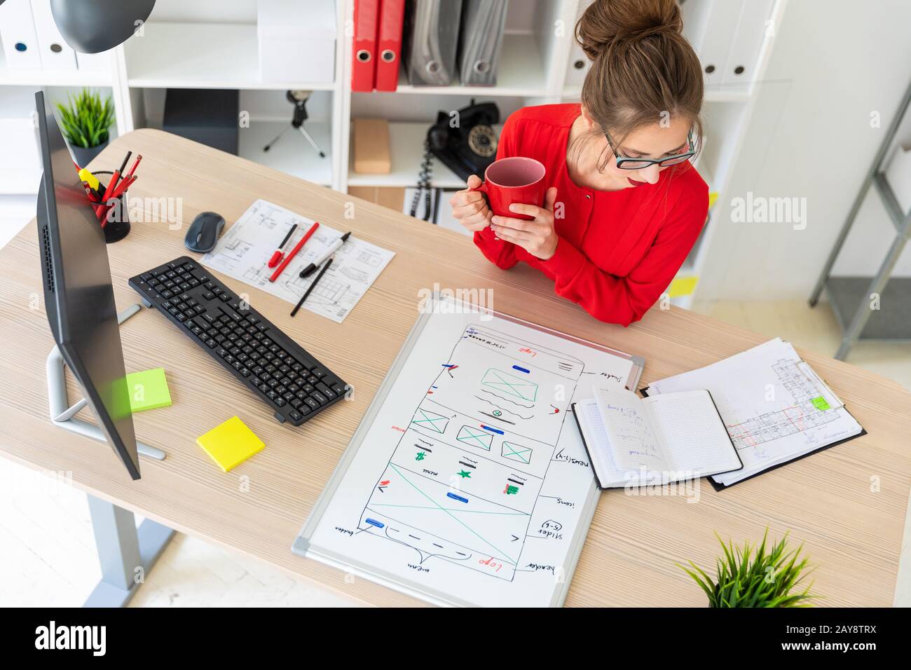 Ein junges Mädchen sitzt am Tisch im Büro, hält einen roten Becher in den Händen und schaut auf den Notizblock. Eine Magnetplatine Stockfoto