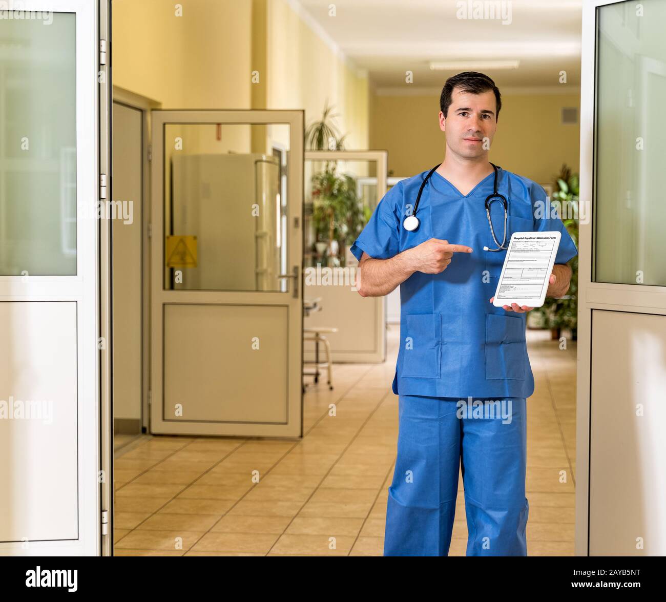 Mittlerer männlicher Arzt in blauen Schrubben, die halten und auf Tablette mit Krankenhauseintrittsformular zeigen Stockfoto