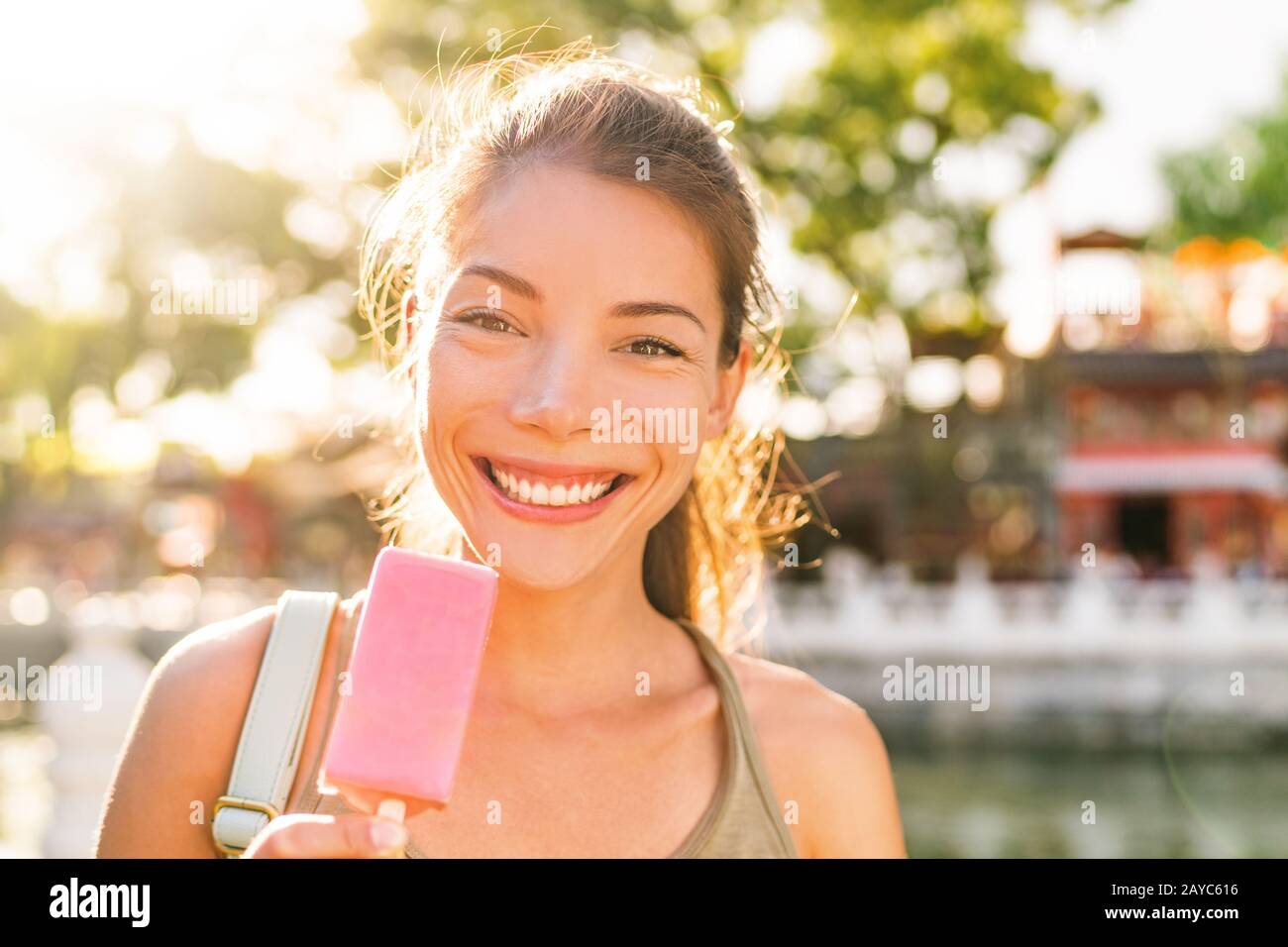 Fröhlicher Sommer verwöhnen Sie asiatisches Mädchen, das Erdbeer-Eis-Stick draußen bei Sonnenuntergang im Sonnenschein isst. Lächelnde Chinesin lächelt am Leben im Stadtpark. Stockfoto