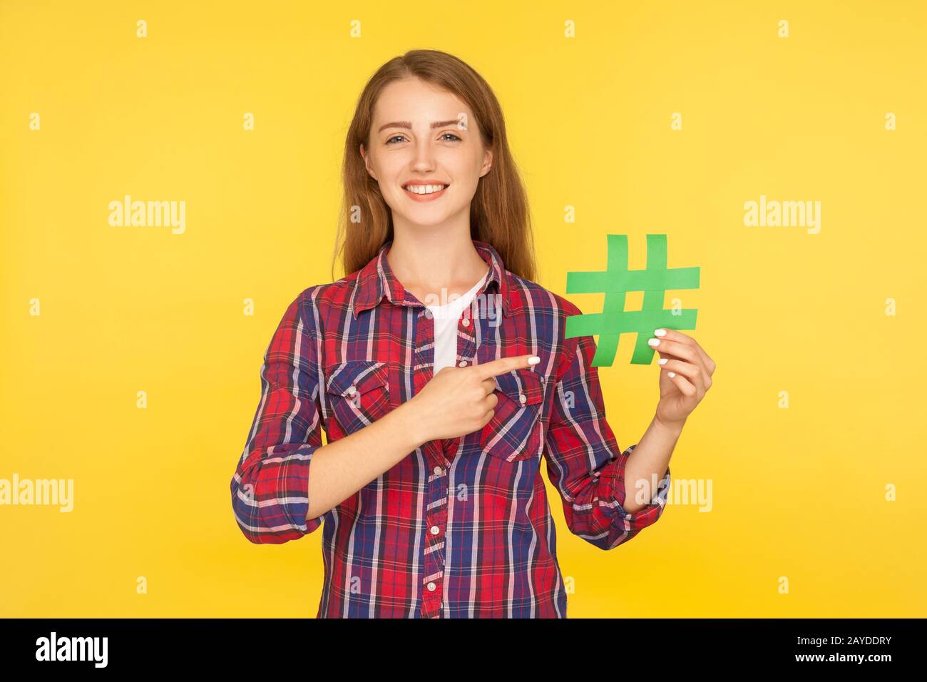 Social Media-Trend, Blogging. Porträt des Ingwermädchens im karierten Hemd, das auf einen grünen Papierhashtag zeigt, Hash-Zeichen zeigt und mit der Kamera lächelt. IND Stockfoto