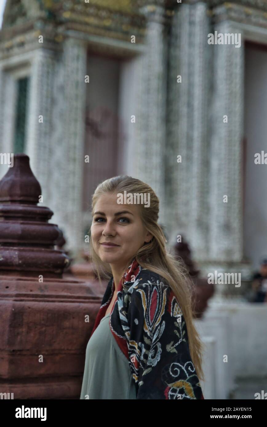 Vertikales Foto eines blonden Mädchens mit dem Haar nach unten, ein Schal, der ihre Schultern bedeckt und ihr Kopf seitlich von einem buddhistischen Tempel in Thailand gedreht wurde Stockfoto