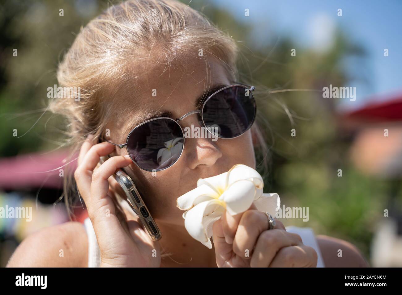 Blondes Mädchen mit Sonnenbrille, das eine weiße Lilie riecht, während sie auf ihrem Handy in Thailand spricht. Reisekonzept Stockfoto