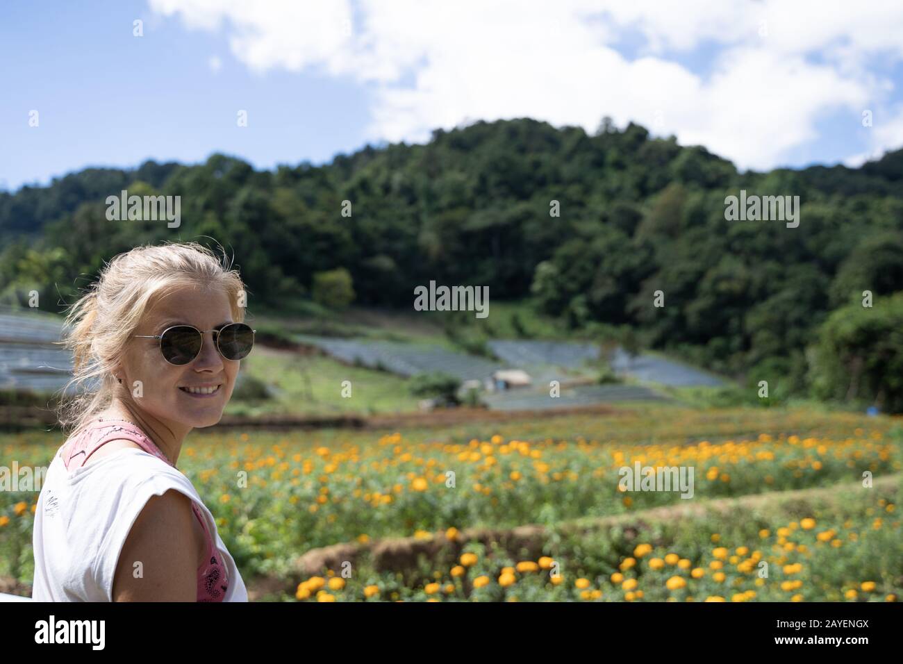 Blondes Mädchen mit Sonnenbrille vor einem Feld gelber Nelken und anderen terrassenförmigen Plantagen mitten im Wald in Thailand Stockfoto