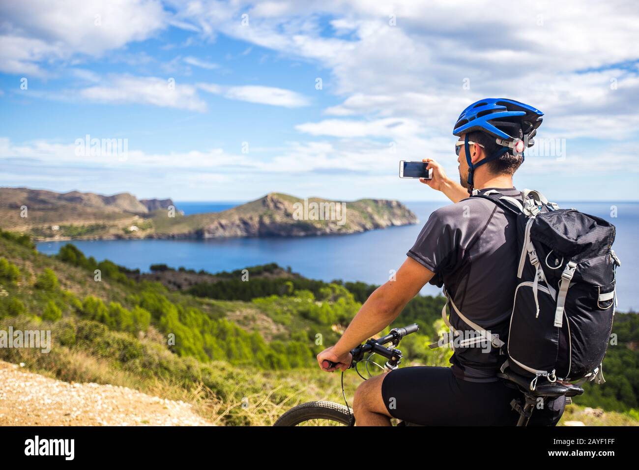 Ein junger Kerl auf einem Mountainbikestrecken in Spanien und macht ein Foto auf einem weißen Telefon im Hintergrund des Mittelmeeres von Stockfoto