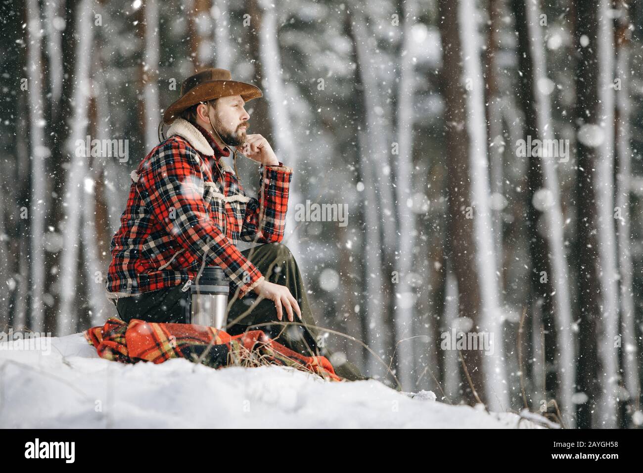 Nachdenklicher männlicher Wanderer in karierter Jacke, der auf einem kleinen verschneiten Waldhügel sitzt und in die Ferne blickt Stockfoto