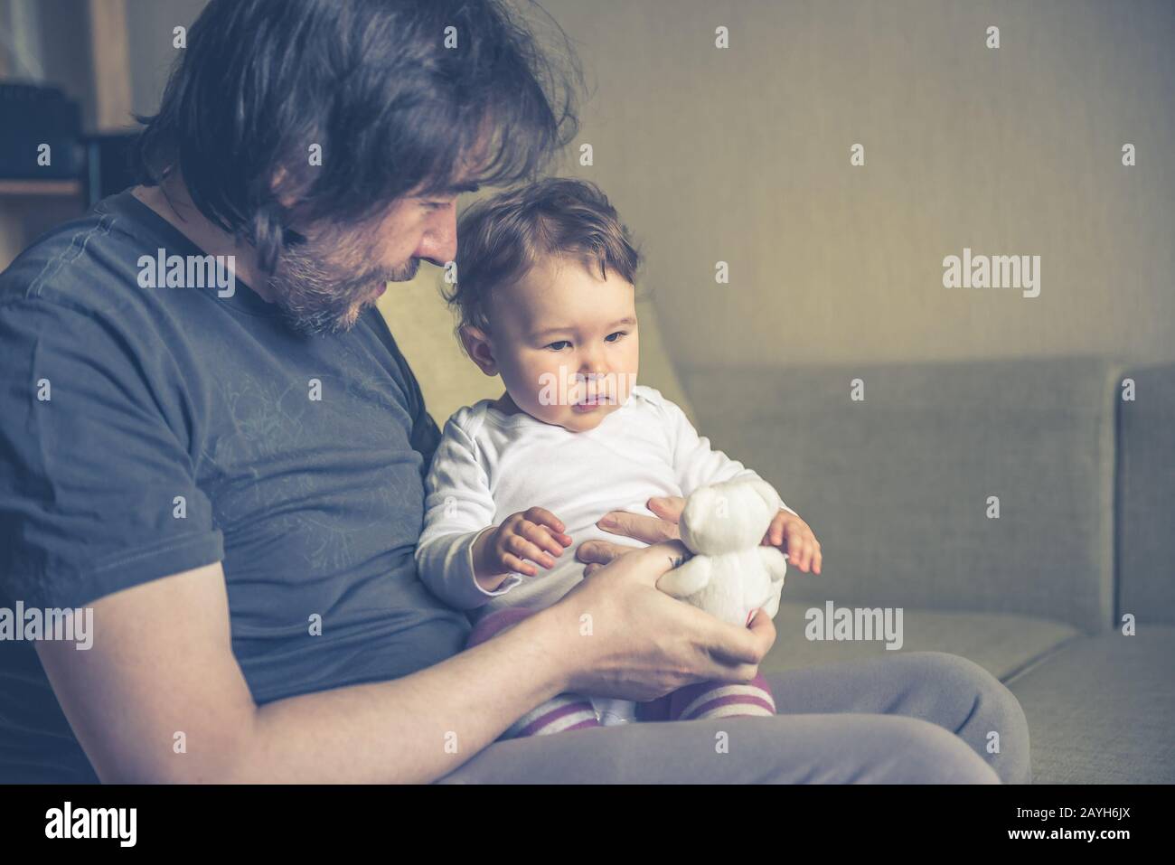 Glücklicher Vater spielt mit seinem Baby zu Hause auf der Couch. Das einjährige Kind sieht sich den Teddybären an. Stockfoto