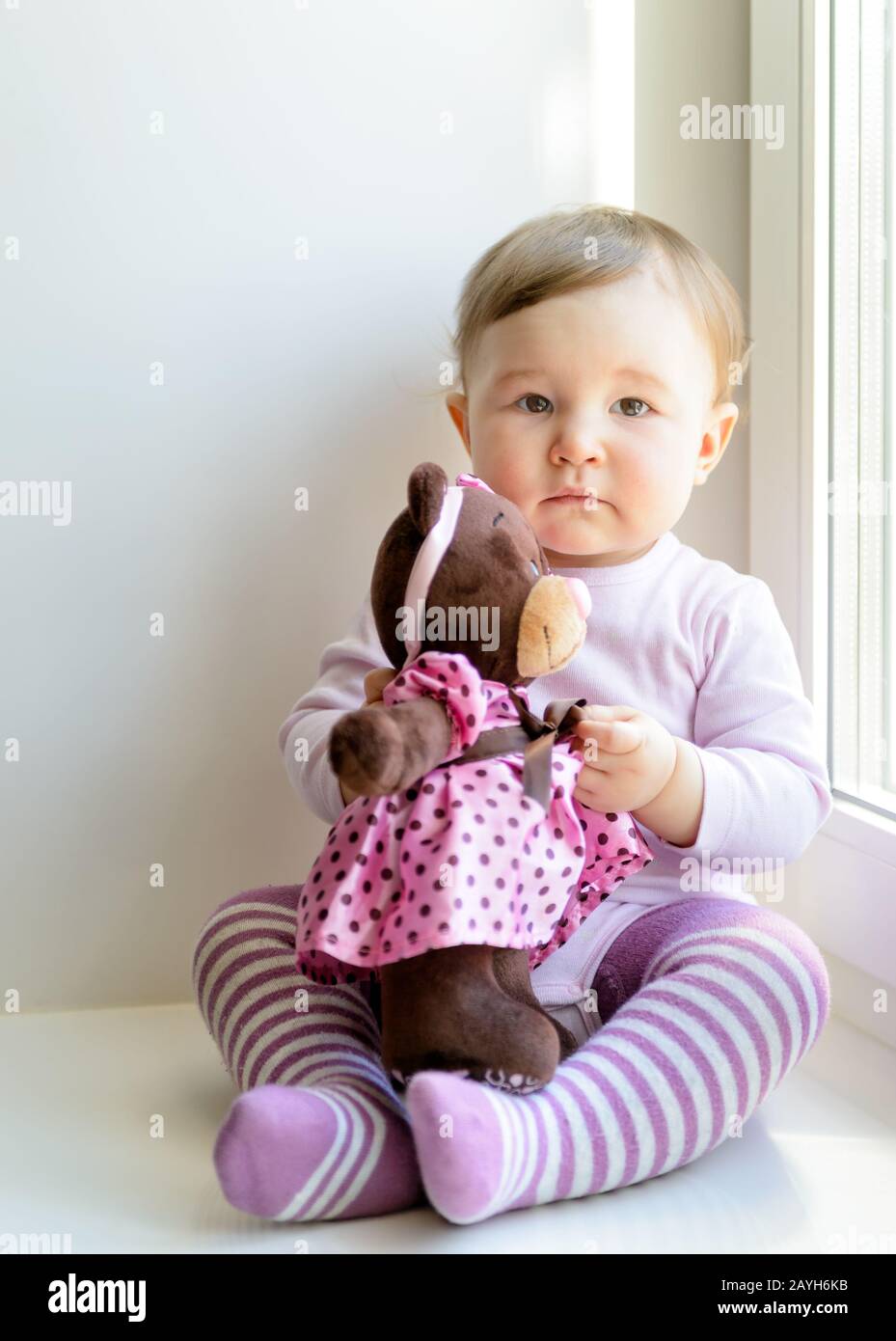 Trauriges nettes Baby-Mädchen, das zu Hause am Fenster mit Spielzeugbären sitzt Stockfoto