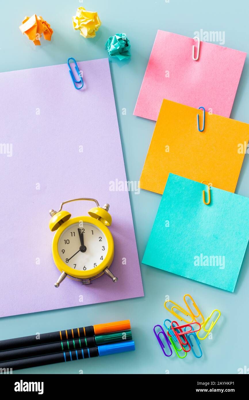 Ein gelber Wecker, sowie bunte, leere Notizkarten. Draufsicht. Bunte Büroklammern, ausgewaschene Papierkugeln, Stifte, Platz für eine Beschriftung Stockfoto