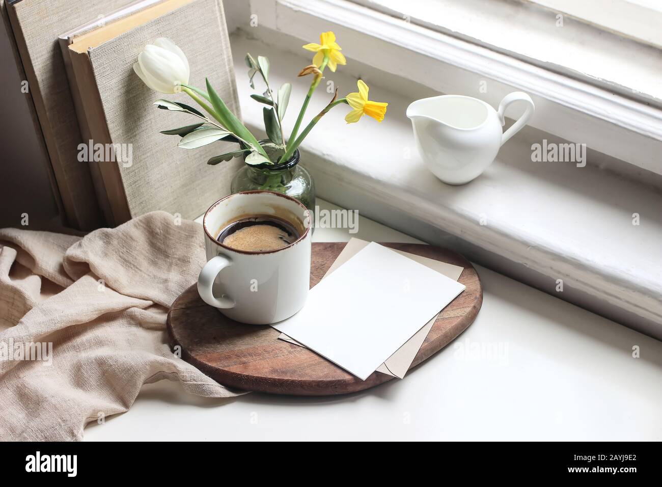 Gemütliches Stillleben im Osterfrühling. Mockup-Szene für Grußkarten. Tasse Kaffee, Bücher, Holzschneidebrett, Milchkrug und Blumenvase auf Fensterbank. Stockfoto
