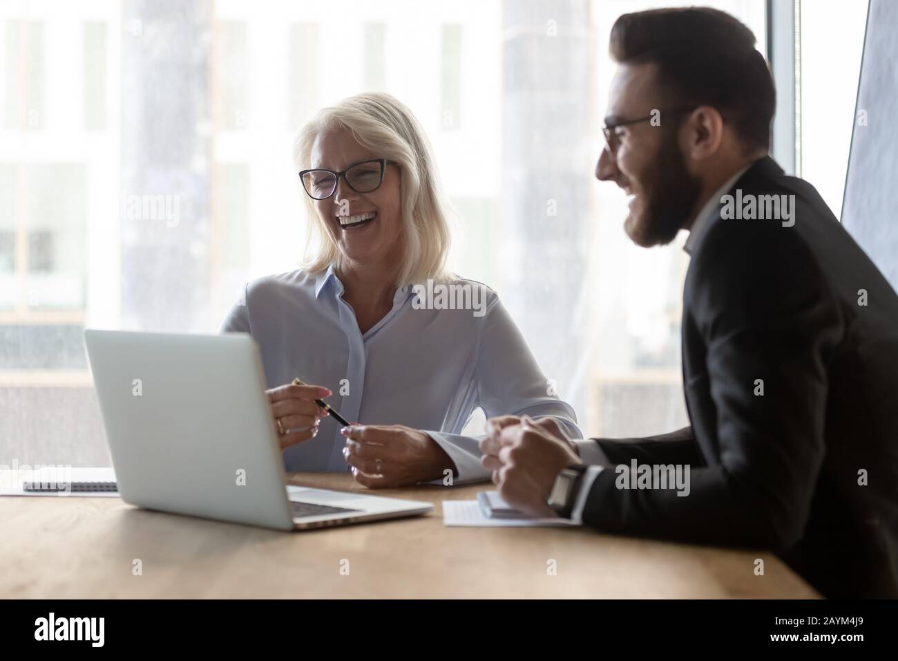 Fröhliche jüngere Kollegin hilft älteren Frauen mit Computeranwendung Stockfoto