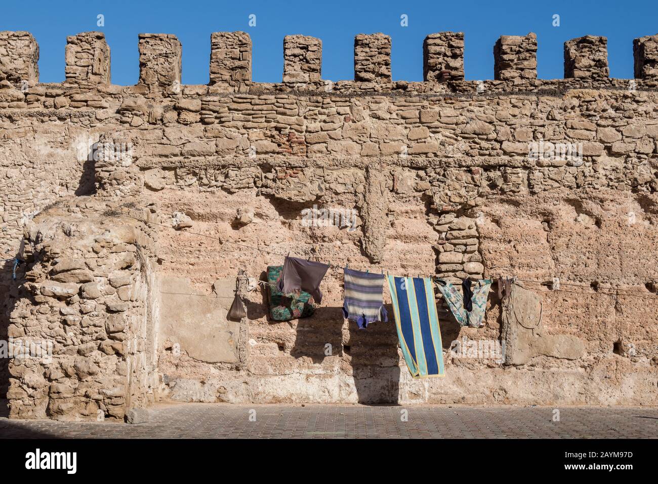 Straße einer Stadt mit einer mittelalterlichen Mauer aus Stein. Seil mit hängender, sauberer Wäsche zum Trocknen in der Sonne. Strahlend blauer Himmel. Medina von Essaouira, Mo Stockfoto
