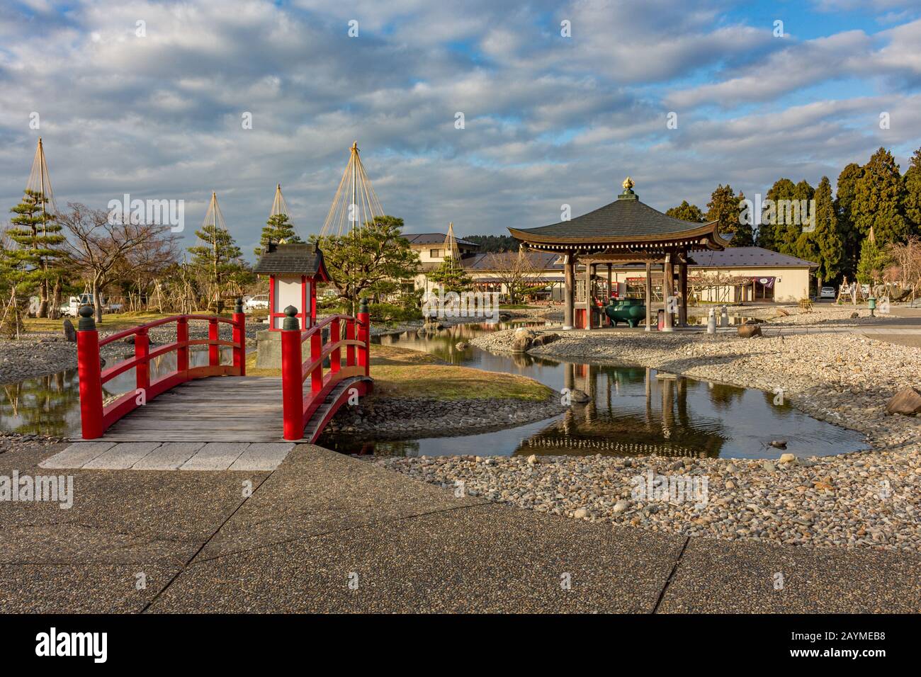 Traditionelle rote Holzbrücke, Teich und Kiefern mit Yukizuri-Seilen, Kurikarafudoji-Tempel, Tsubata, Japan. Stockfoto