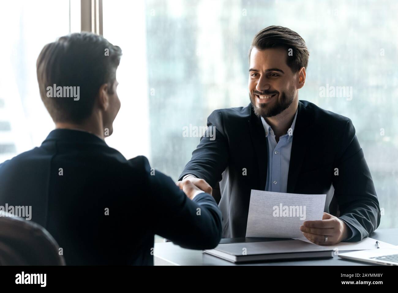 Lächelnder bärtiger Geschäftsmann im Anzug schüttelt mit Arbeitssuchenden die Hände. Stockfoto