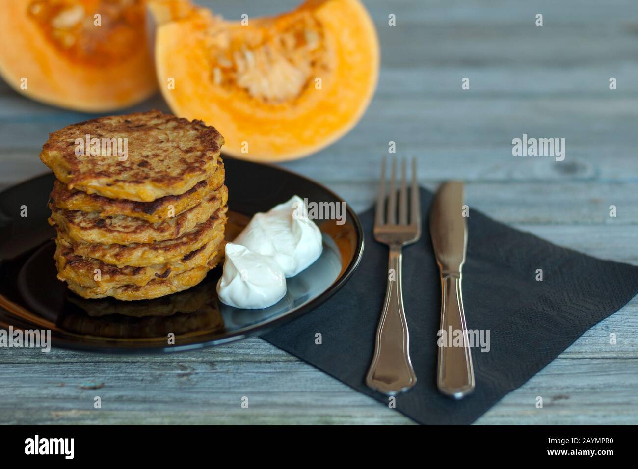Pfannkuchen mit natürlichem Yougurt auf schwarzem Teller, Gabel, Messer, PapierServiette serviert auf grauem Holztisch, geschnittene Stücke und orangefarbener Kürbis auf dem Hintergrund Stockfoto