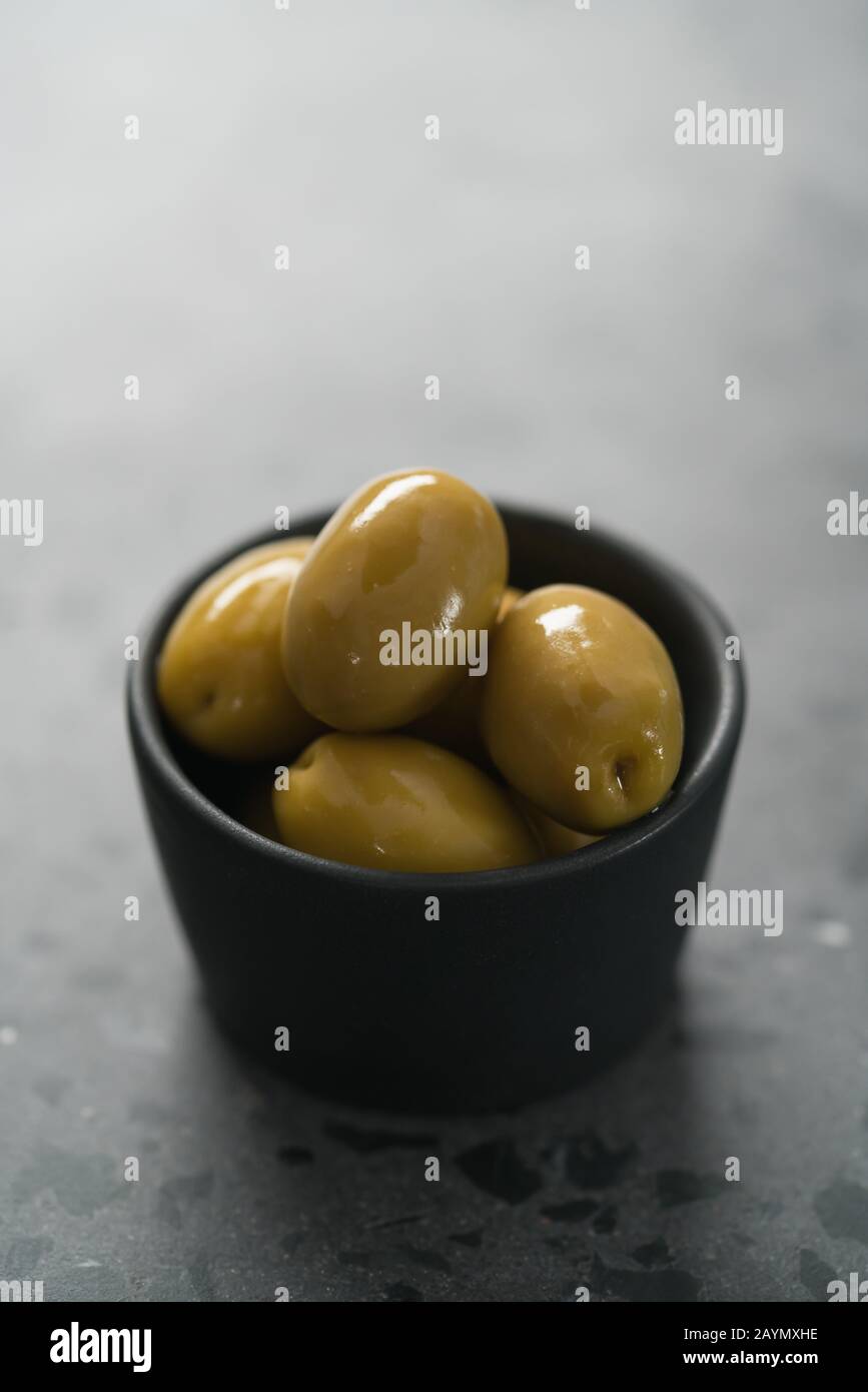Ganze große grüne Oliven in kleiner schwarzer Schüssel auf Betongrund, flacher Fokus Stockfoto