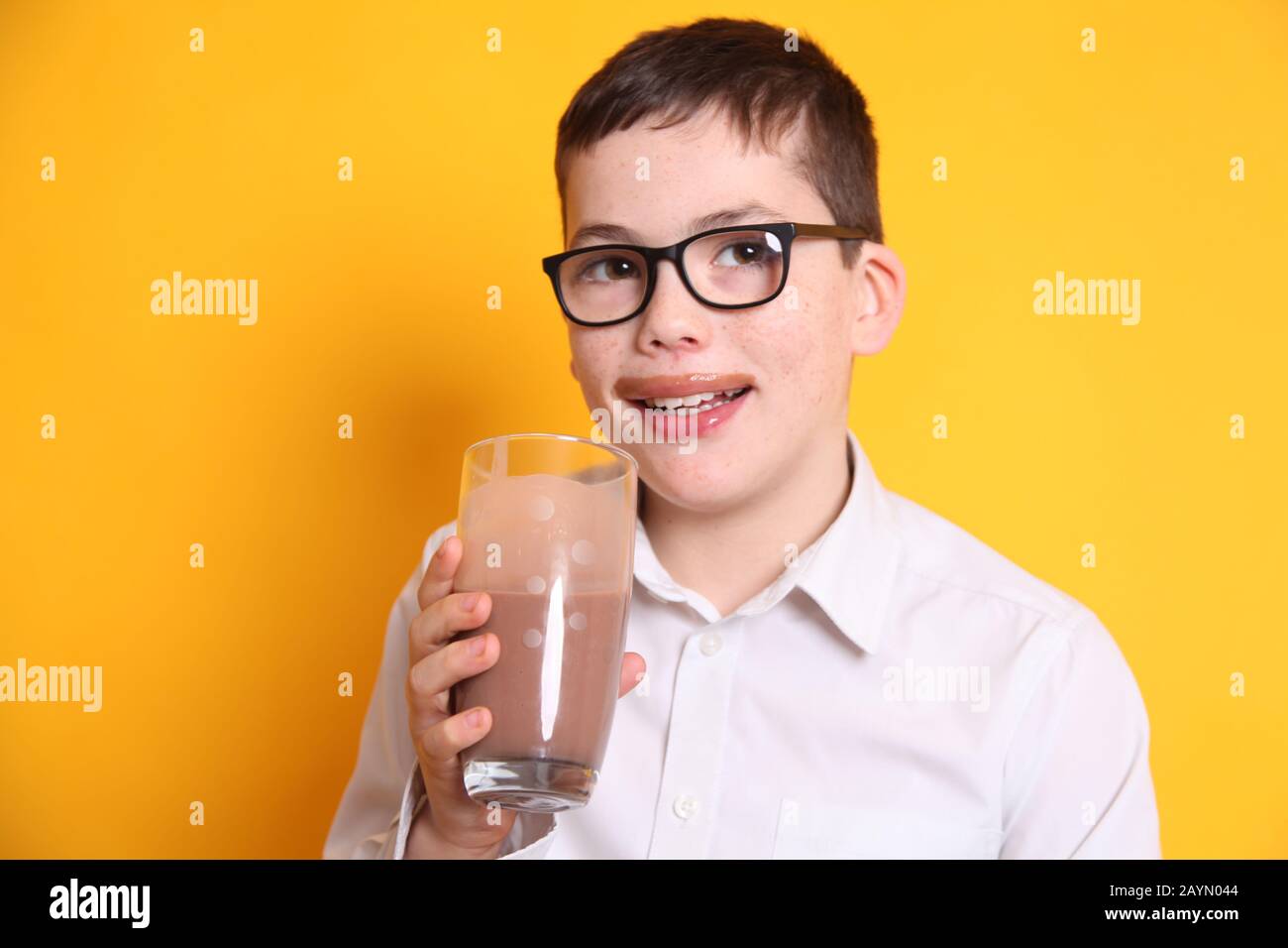 Ein junger Junge, der eine Brille im Alter von 8 Jahren trägt, trinkt ein Glas Schokoladenmilch mit der Oberlippe, die auf gelbem Hintergrund mit Schokolade bedeckt ist Stockfoto