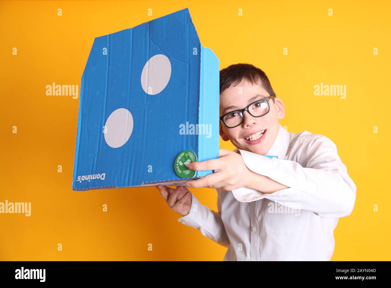 Ein kleiner Junge mit einer Brille im Alter von 12 Jahren und einer großen Domino's Pizza Box auf gelbem Hintergrund Stockfoto