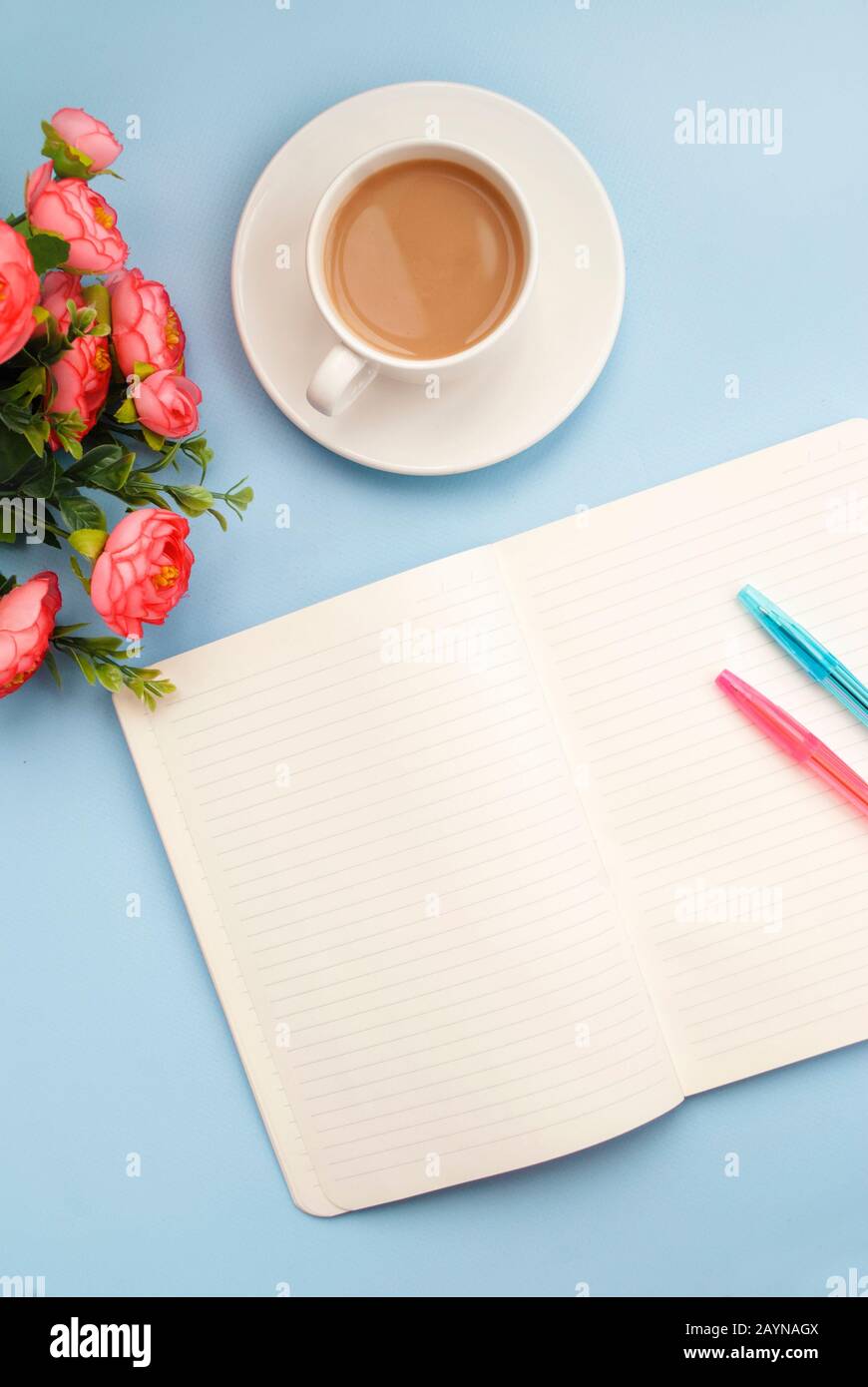 Konzept Arbeitsplatz. Weiße Tasse Kaffee mit Milch, weißes, offenes Notizbuch für Notizen, Kugelschreiber, pinke Blume auf blauem Hintergrund, Platz für Text, Kopie Stockfoto