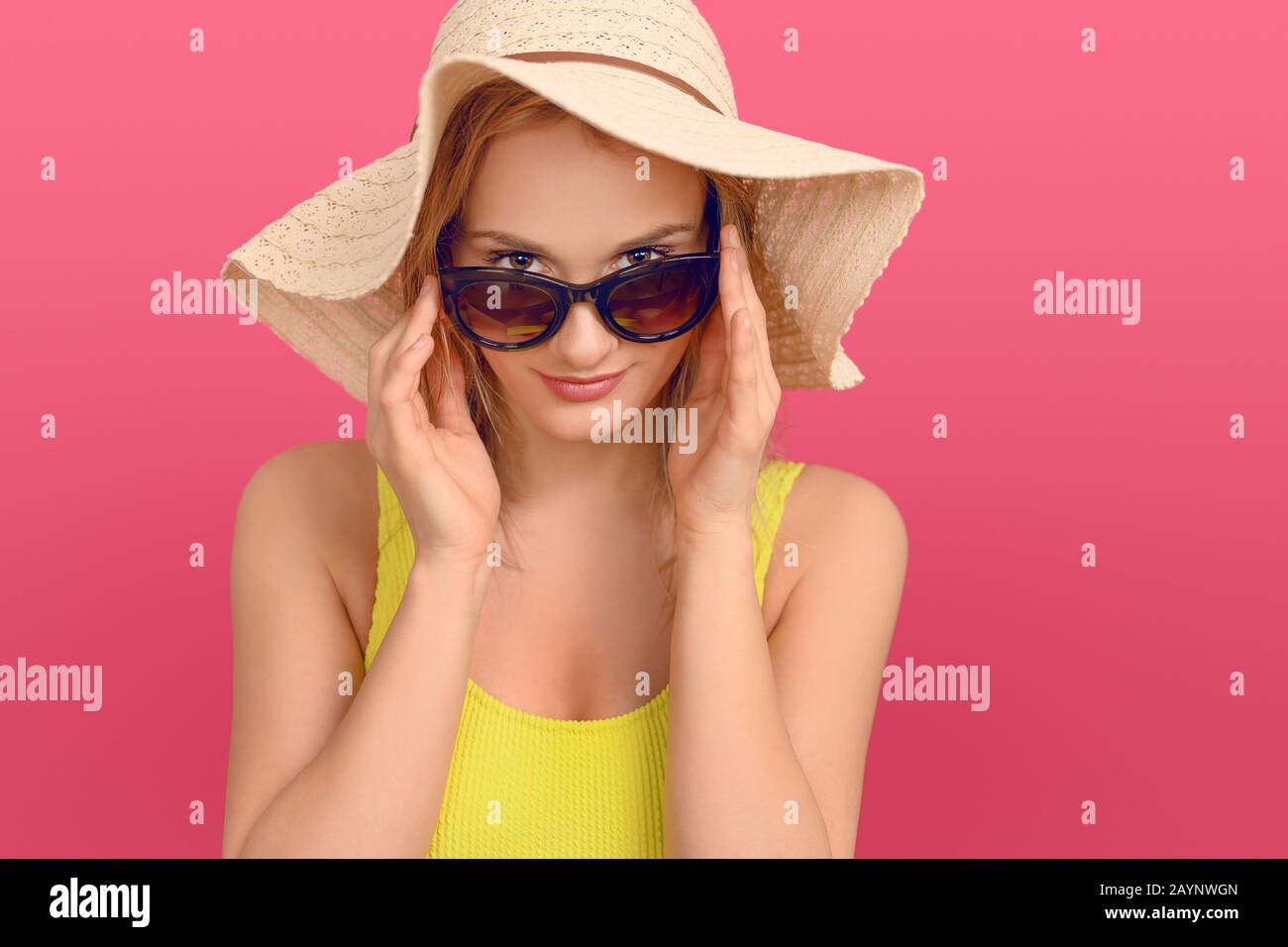 Attraktive, schicke junge Frau im Sommer-Outfit mit einem großen Floppy-Sonnenhut, der mit einem freundlichen Lächeln über die Oberseite ihrer Sonnenbrille an der Kamera schält Stockfoto