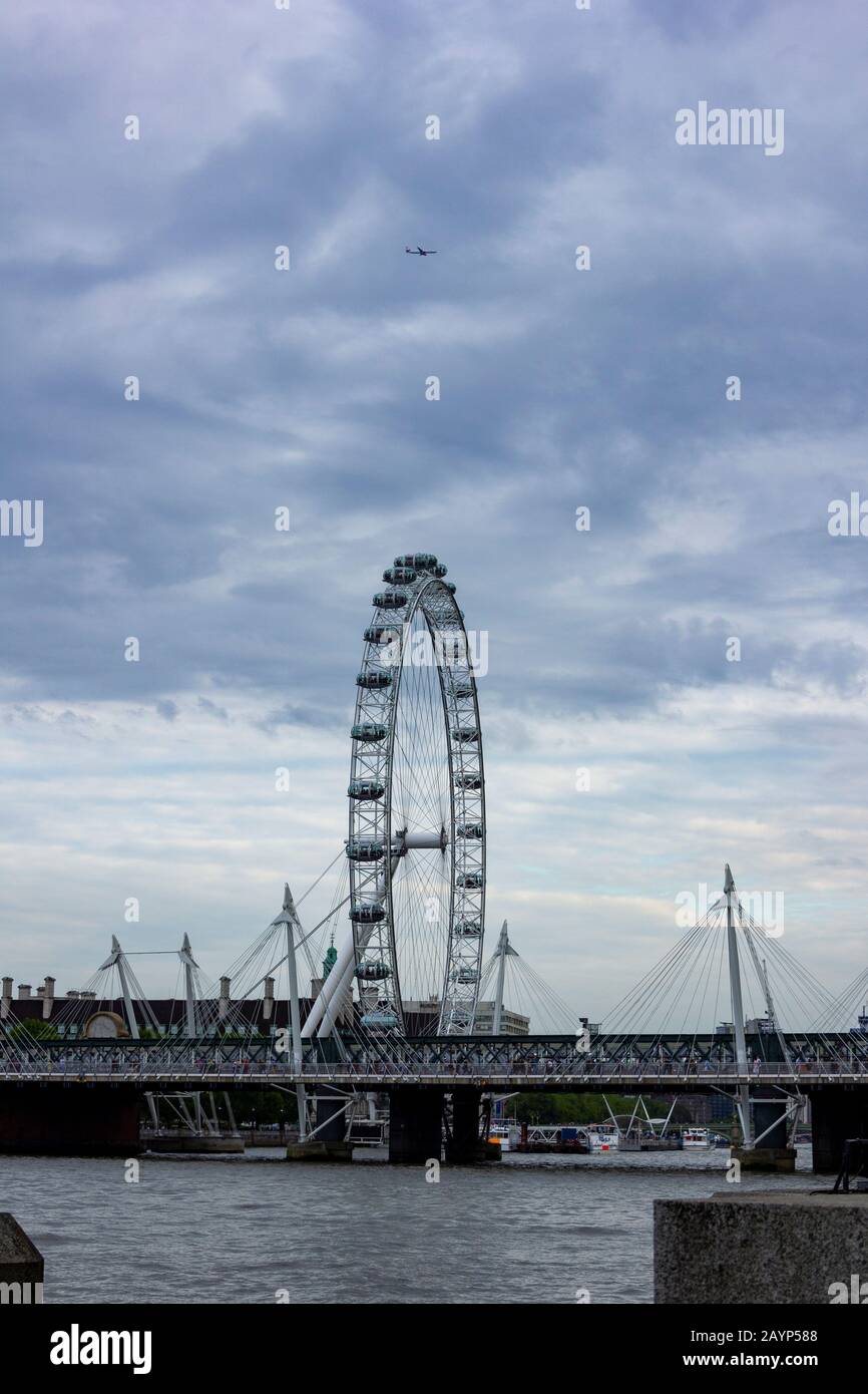 London/England - 24. Mai 2019: Ein Porträt des London Eye mit einem stimmungsvollen Himmel und einem Flugzeug darüber und einer Brücke über die Themse. Stockfoto