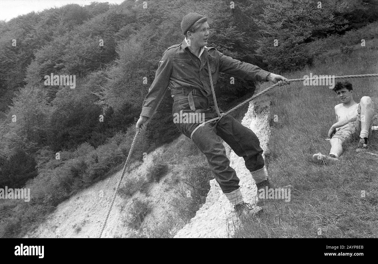 1950er Jahre, historisch, an der Spitze einer steilen Klippe, ein junger Mann in Jeans und Kletterschuhen, an einem Seil haltend, um seinen Körper gewickelt, kurz davor, sich abzuseißen oder die steile, zerklüftete Steigung hinunter zu steigen, England, Großbritannien. Sein Freund beobachtete ihn entspannend auf dem grasbewachsenen Ufer nahe der Klippe. Stockfoto