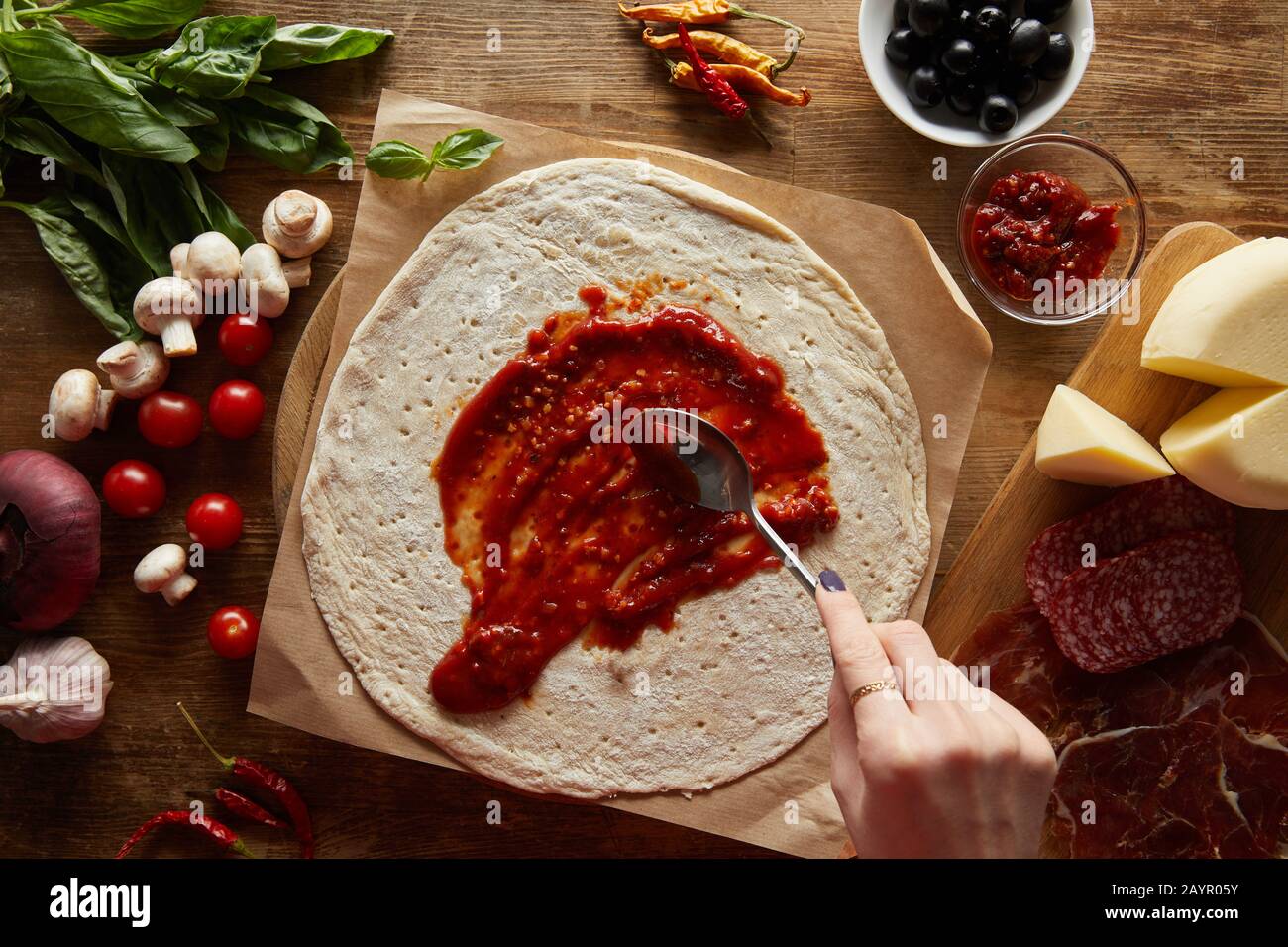 3/4-Ansicht der Frau, die Tomatensauce auf Teig in der Nähe von Zutaten für Pizza auf Holztisch zufügt Stockfoto