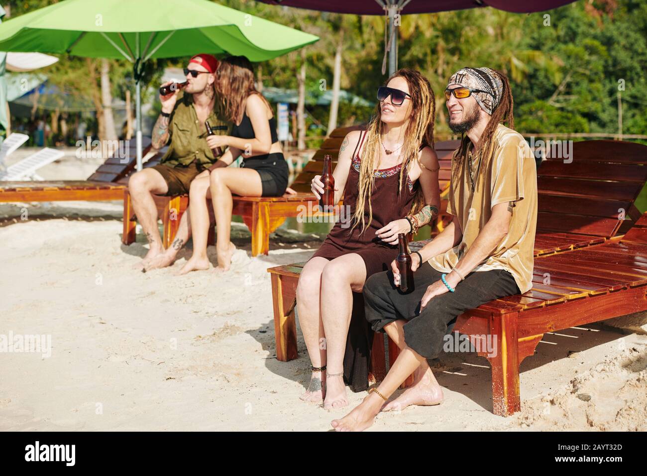 Positive Paare sitzen auf Holz-Chaise-Lounges am Strand, trinken Bier und genießen eine schöne Aussicht Stockfoto
