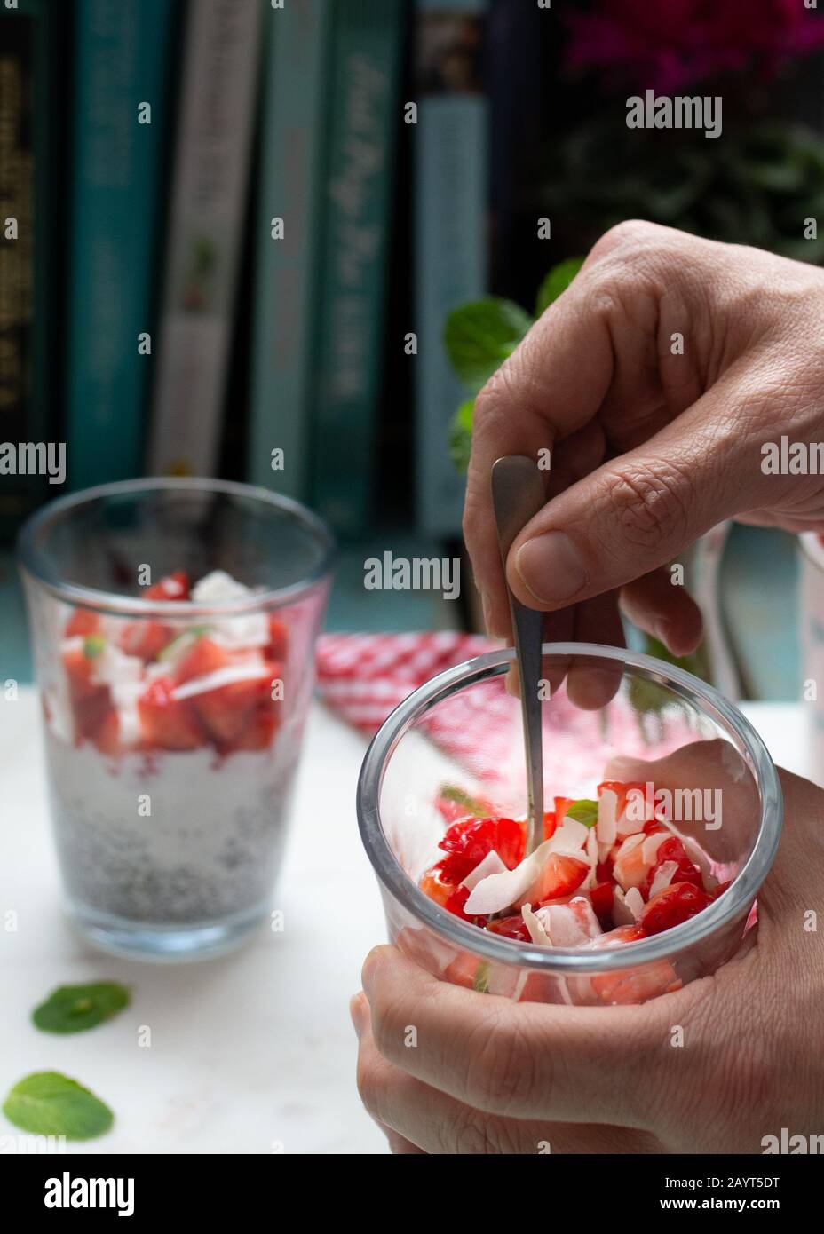 Man hält einen Löffel in ein hausgemachtes Glas voller Erdbeeren mit chia, Cocochips, Milch und Minzblättern. Stockfoto