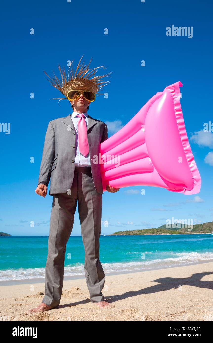 Ein ernsthafter Geschäftsmann, der Strohhut und eine überdimensionale Sonnenbrille trägt, die mit einem rosafarbenen aufblasbaren LILO Floß an einem tropischen Strand steht Stockfoto