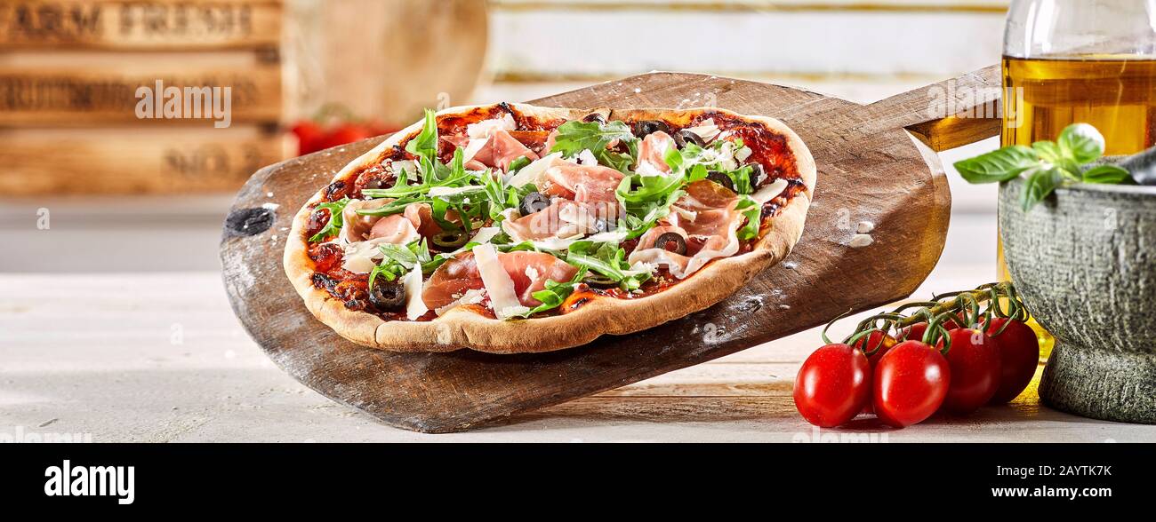 Zerknustete italienische Prosciutto Pizza mit frischer Rakete und Parmesan Käsebesatz auf einem alten Holzpaddel mit Zutaten in einer Pizzeria in a Stockfoto