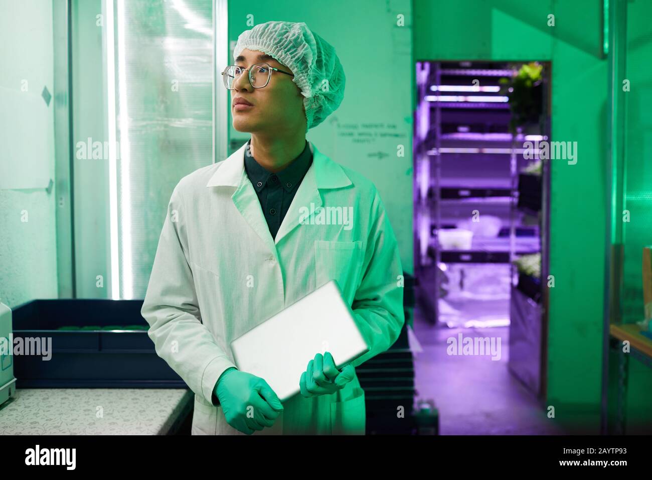 Taille oben Porträt junger asiatischer Arbeiter, die Schutzkleidung tragen, die im Biolabor steht, das von grünem Licht beleuchtet wird, Kopierraum Stockfoto