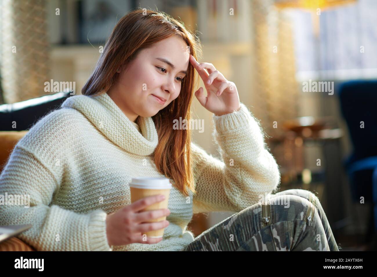 Fröhliches modernes Teenager-Mädchen mit roten Haaren im weißen Pullover, das in der Nähe der Couch sitzt und im modernen Wohnzimmer am sonnigen Wintertag eine Tasse heißes Getränk serviert. Stockfoto