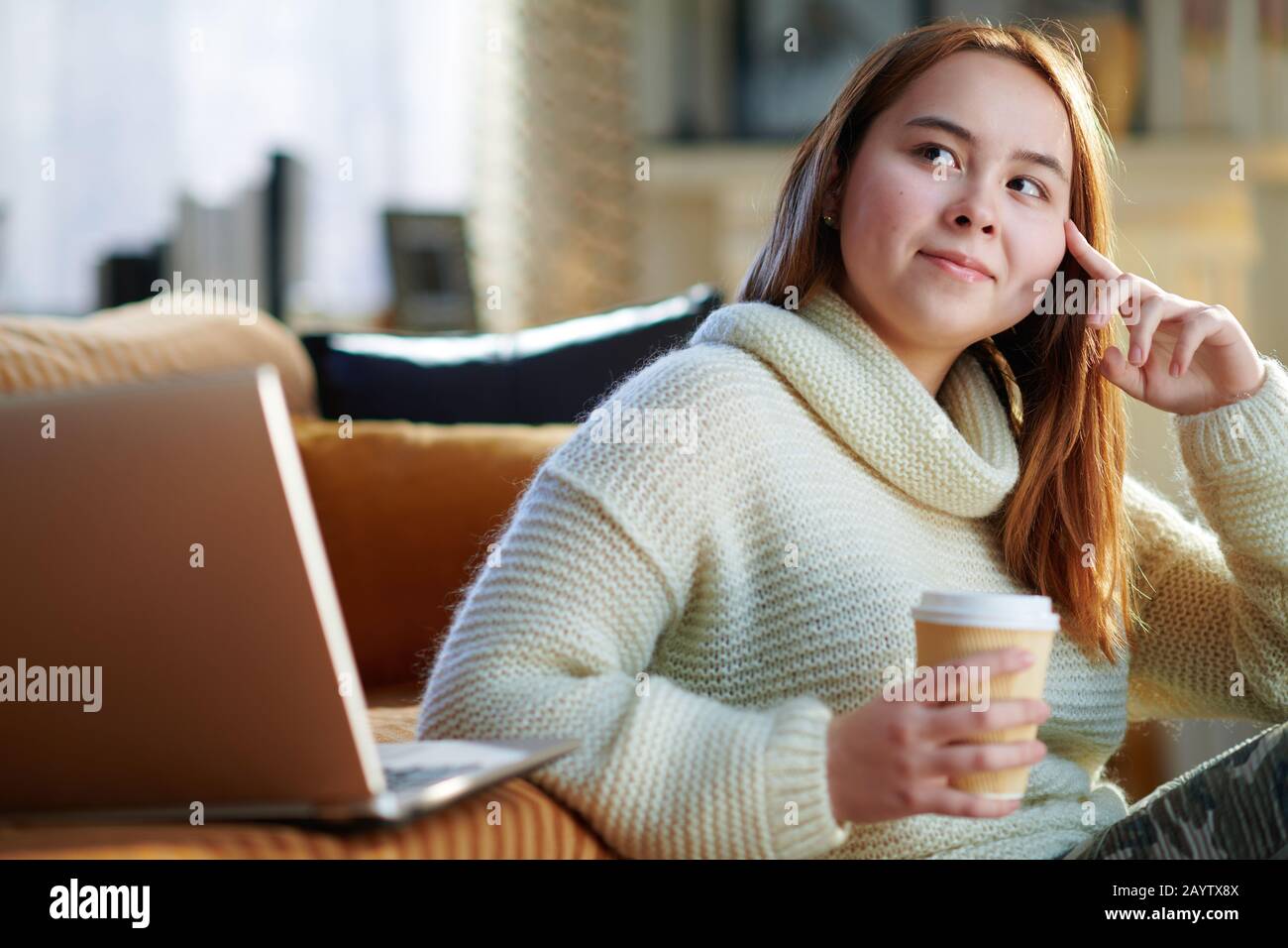Modernes Mädchen mit roten Haaren in weißem Pullover, das in der Nähe der Couch sitzt und im modernen Wohnzimmer in sonniger Sonne eine Tasse heißen Getränks und Laptop hat Stockfoto