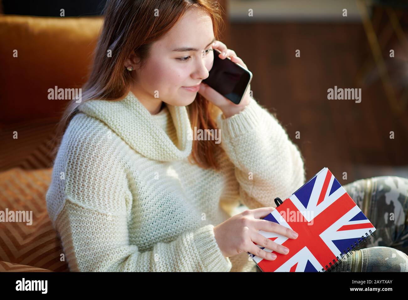 Fröhliches Mädchen der modernen Jugend mit roten Haaren in weißem Pullover mit Notizbuchfarben britischer Flagge, das auf einem Handy im modernen Haus in sonniger Lage spricht Stockfoto