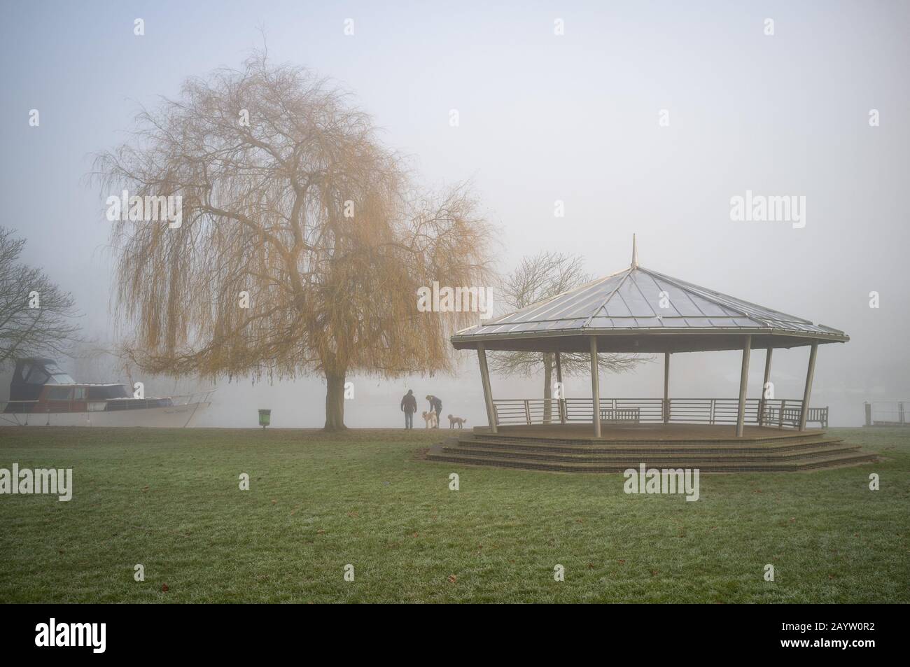 Hundegänger treffen sich am frühen Morgen auf dem Schlepppfad in Nebel von einem Weidenbaum und einem vermoorten Kabinenkreuzer Stockfoto