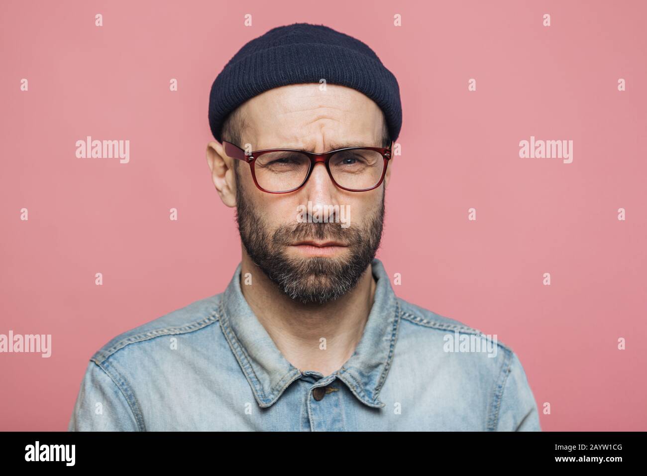 Unglücklich Unzufriedenheit unrasierten Mann schaut mit mürrischen Ausdruck in Kamera, trägt eine Brille schwarzen Hut und stilvolle Jeanshemd, über Rosa backgro isoliert Stockfoto