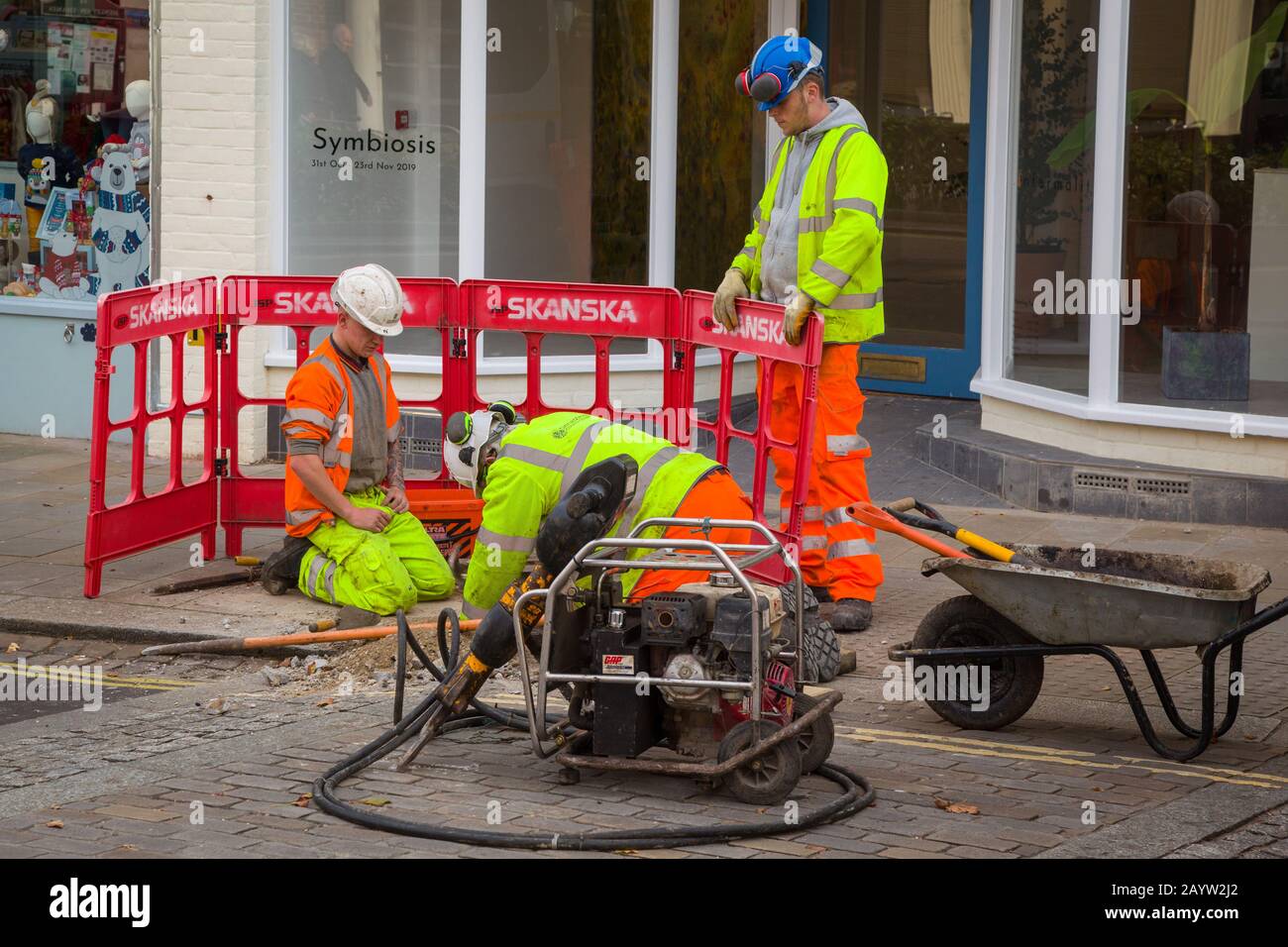Arbeiter in gelben fluoreszierenden Jacken und Harthüten, die ein Loch im Bürgersteig für den Bauunternehmer Skanska mit roten Skanska-Barrieren behüten Stockfoto