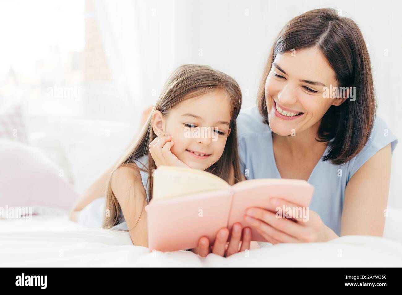 Hübsche Frauen Mutter und ihre kleine Tochter lesen Sie interessantes Buch im Schlafzimmer, auf comfortabled Bett liegen während der Morgen. Schöne Mama liest Märchen für fairty Stockfoto