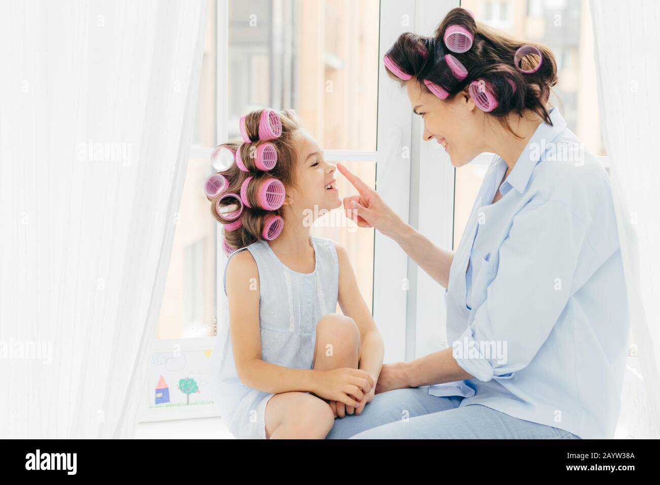 Horizontale Schuß von der hübschen jungen Mutter und Tochter mit Lockenwicklern im Haar, in gute Laune, genießen Sie Zeit zu Hause, für den Urlaub vorzubereiten. Happy Family co Stockfoto