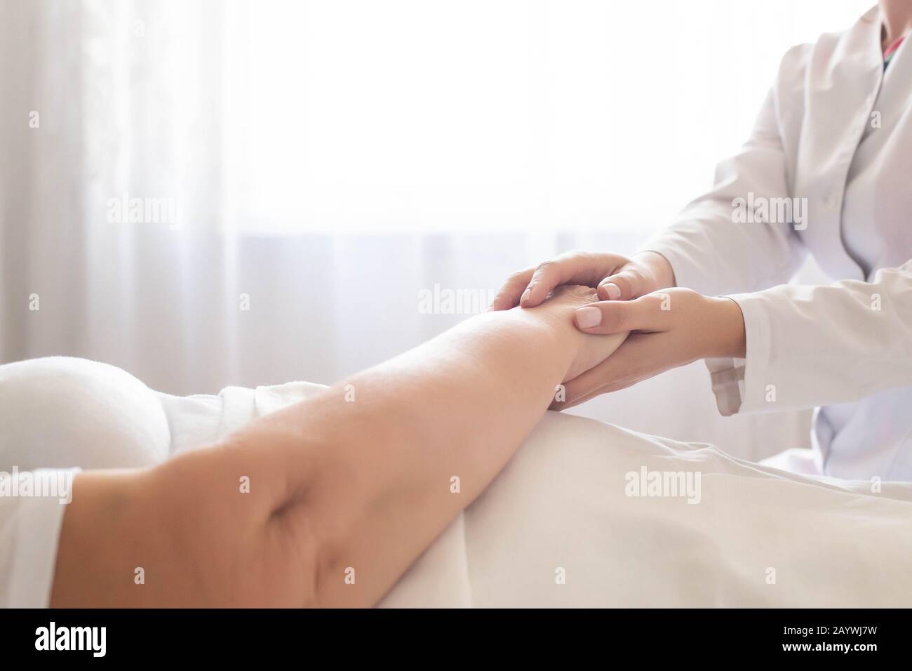 Der Arzt hält die Hand eines Patienten einer älteren Frau auf einem Hintergrund eines Fensters. Konzept der hygienischen Patientenversorgung in einem Krankenhaus, Geriatrie Stockfoto